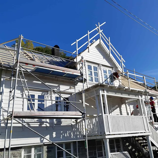 Et hvitt trehus med stillas og arbeidere som utfører takrenovering.